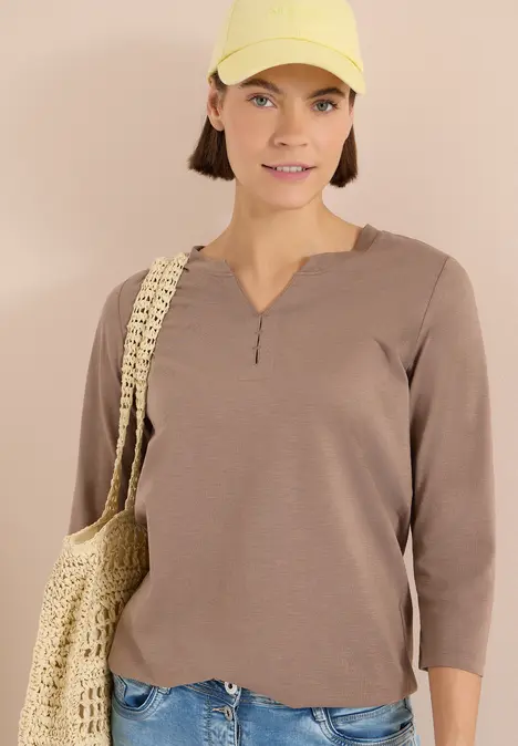 Person trägt beigefarbenes Langarmshirt und geflochtene Tasche vor neutralem Hintergrund.