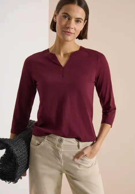 Frau in burgunderfarbenem Dreiviertelarmshirt mit V-Ausschnitt, beiger Hose und schwarzem, geflochtenem Korb.
