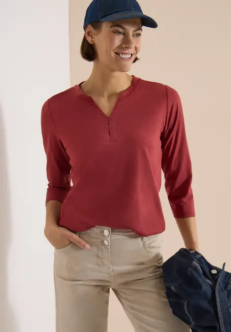 Frau trägt rotes Langarmshirt mit Knopfleiste und helle Hose, hält lässig eine Jacke in der Hand.