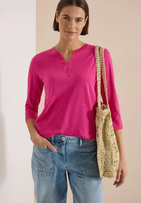 Frau trägt pinkes 3/4-Arm-Shirt, blaue Jeans und hält eine gehäkelte Strohtasche.