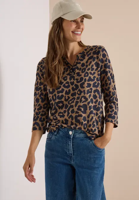 Lächelnde Frau mit beige Cap, Leo-Print-Shirt und blauen Jeans, Hand in Tasche, vor hellem Hintergrund.