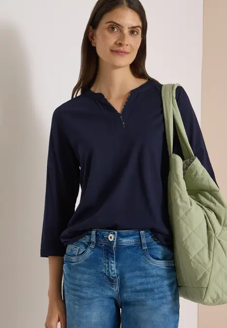 Eine Frau trägt ein dunkelblaues Langarmshirt, Jeans und eine grüne gesteppte Tasche lässig über der Schulter.