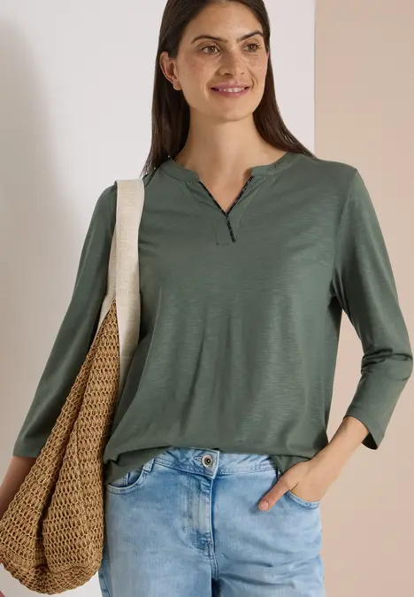 Frau in grünem Longsleeve, trägt Jeans und Korbtasche, lächelt vor hellem Hintergrund.
