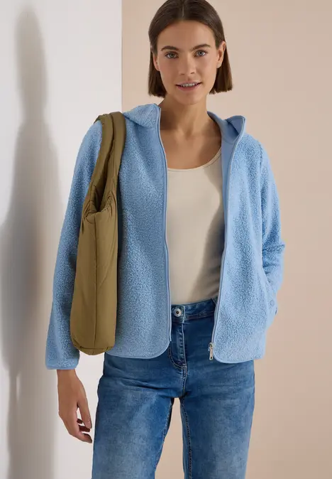 Frau trägt hellblauen Fleece-Pullover mit Kapuze, weißes Top, Jeans und olivgrüne Umhängetasche.