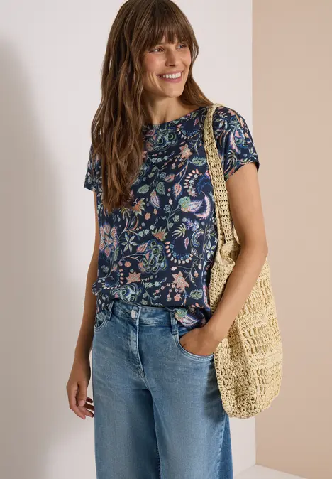 Vrouw in bloes, spijkerbroek en beige gehaakte tas, glimlachend.