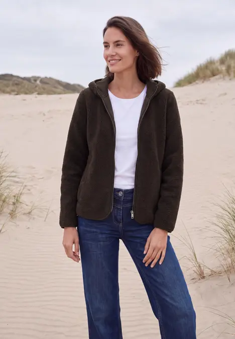 Frau in dunkelgrüner Fleecejacke, weißem T-Shirt und blauen Jeans steht lächelnd am Dünenstrand