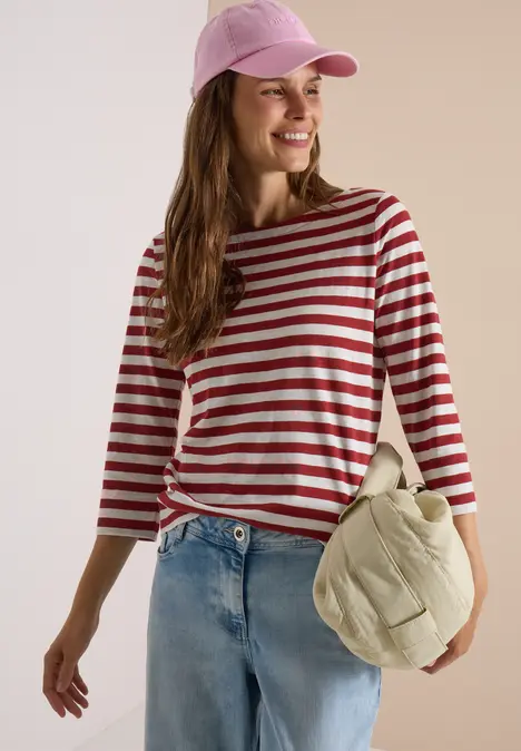 Frau in rot-weiß gestreiftem Shirt, Jeans und rosa Baseballcap trägt lässige Tasche und lächelt.