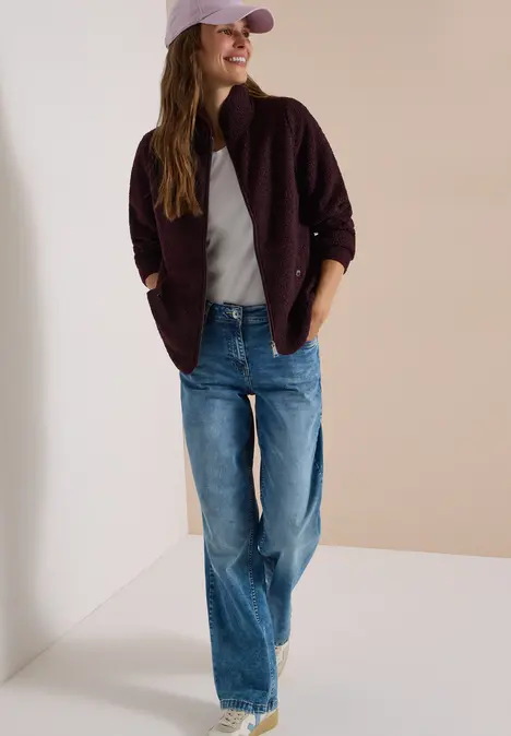 Frau in Jeans, grauem T-Shirt, rosa Cap und dunkelrotem Cardigan steht lässig vor einer Wand.