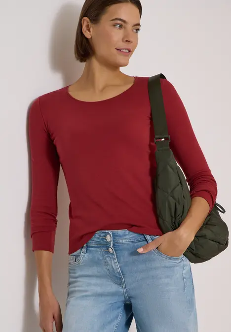 Frau in rotem 3/4-Arm-Shirt, blauer Jeans und grüner Stepp-Umhängetasche vor hellem Hintergrund.