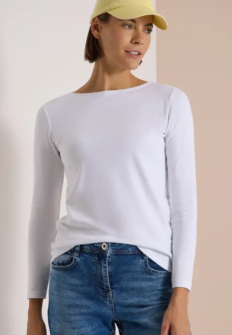 Frau trägt weißes Langarmshirt, blaue Jeans und gelbe Cap, steht lässig vor hellem Hintergrund.