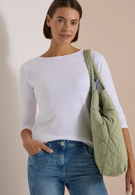 Frau in weißem Langarmshirt, blauen Jeans und gesteppter grüner Umhängetasche.
