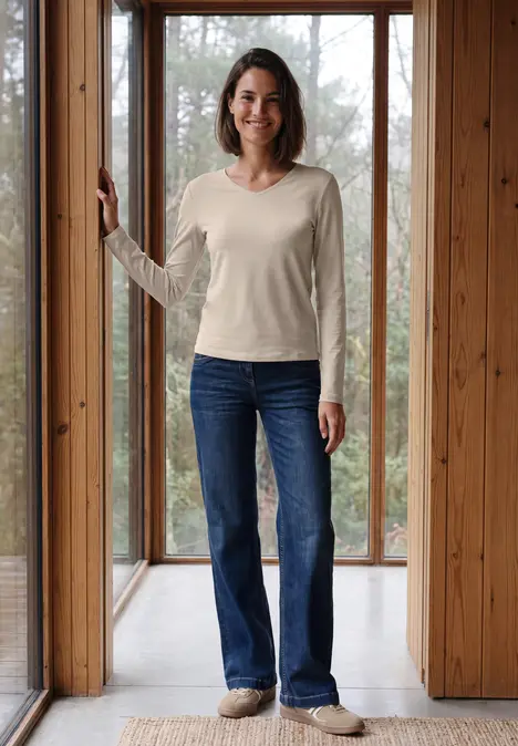 Eine lächelnde Frau in hellem Pullover und Jeans steht vor bodentiefen Holzfenstern.