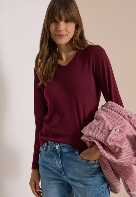 Frau in Jeans und weinrotem Langarmshirt mit braunen Haaren hält rosa Jacke und lächelt.