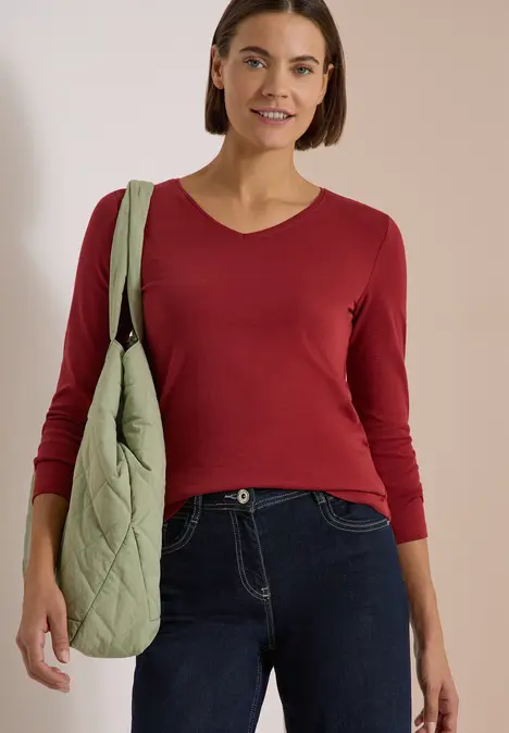 Eine Frau trägt ein rotes Langarmshirt, dunkle Jeans und eine grüne gesteppte Tasche.