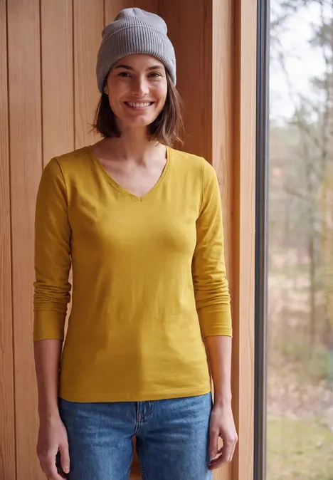 Junge Frau mit gelbem Langarmshirt, blauer Jeans und grauer Mütze steht lächelnd am Hauseingang.
