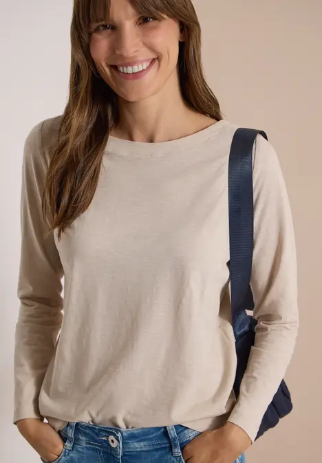Junge Frau lächelt, trägt beige Langarmshirt und Rucksack, steht vor hellem Hintergrund.