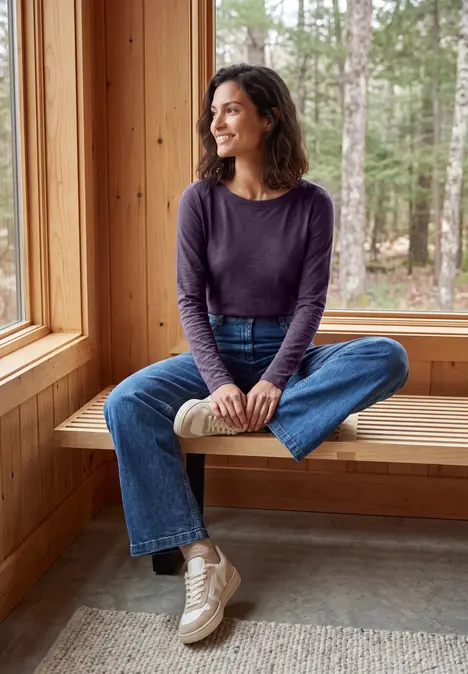 Eine lächelnde Frau in Jeans und langärmligem Shirt sitzt auf einer Holzbank am Fenster in einer Hütte.
