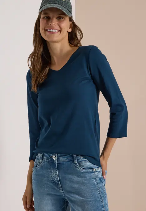Frau in blaugrünem 3/4-Arm-Shirt, Jeans und Cap lächelt.