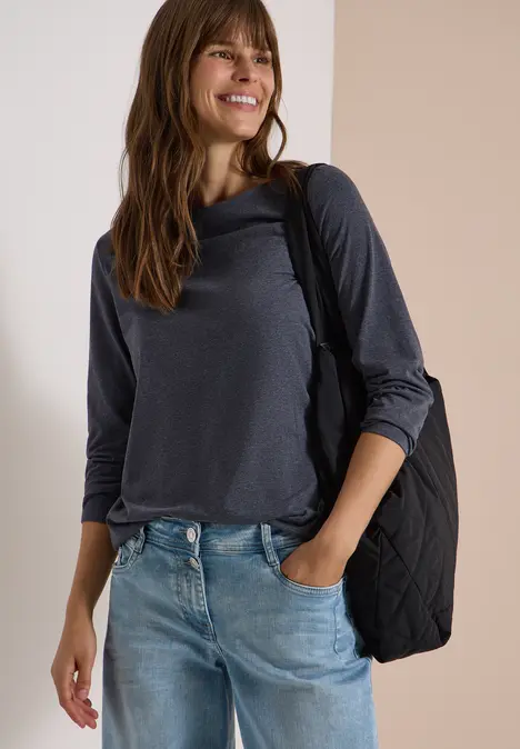 Junge Frau in lässigem grauen Langarmshirt, Jeans und Schultertasche, lächelnd an einer hellen Wand.
