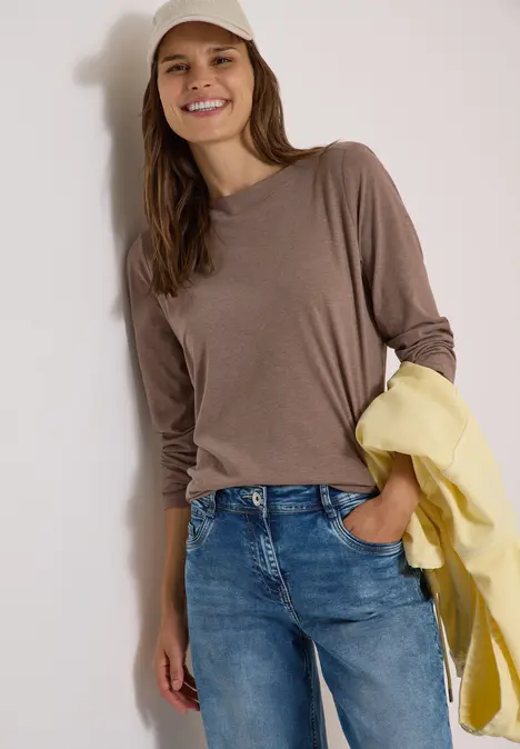 Junge Frau lächelt in braunem Langarmshirt, Jeans und weißer Kappe, hält eine gelbe Jacke.