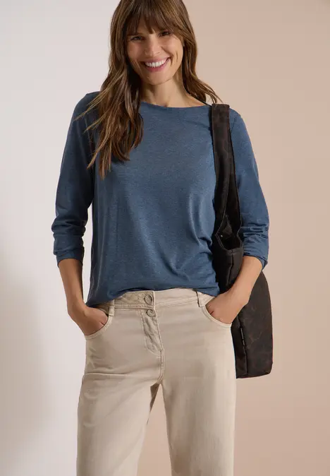 Frau in beiger Hose und blauem Langarmshirt, lächelnd, Hände in den Taschen, mit schwarzer Umhängetasche.