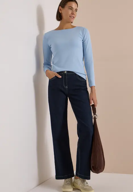 Frau steht mit hellblauem Langarmshirt, weiten dunklen Jeans und brauner Tasche vor einer Wand.