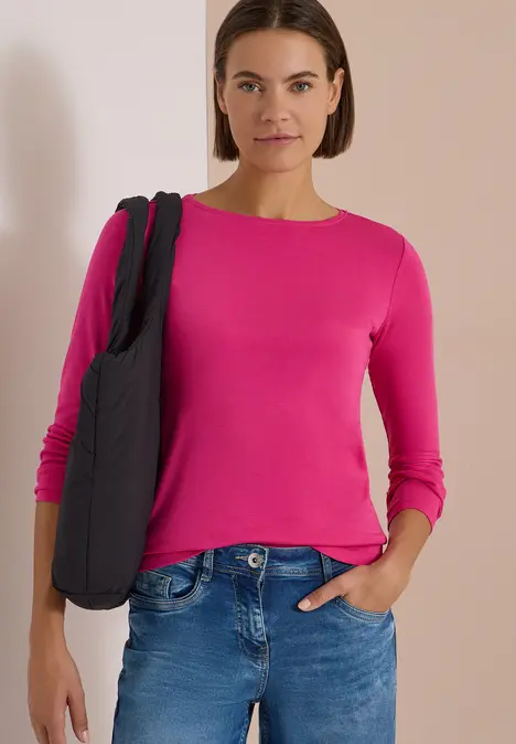 Frau mit pinkem Langarmshirt und Jeans trägt eine schwarze Tasche und steht vor hellem Hintergrund.