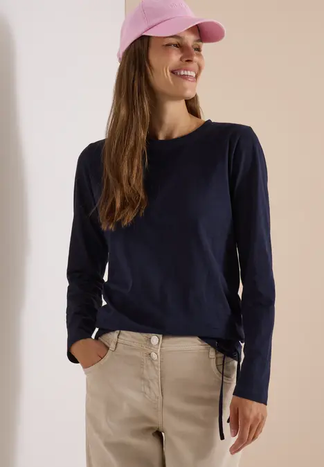 Eine junge Frau lächelt, trägt dunkelblaues Langarmshirt, beige Hose und rosa Cap.
