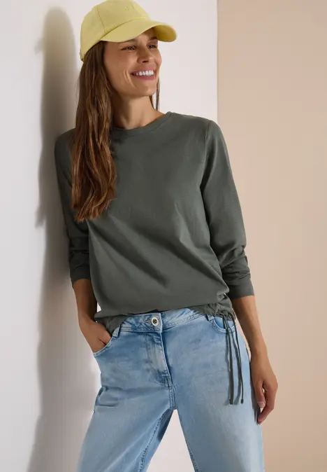 Eine lächelnde Frau trägt ein grünes Langarmshirt, helle Jeans und eine gelbe Baseballkappe, lehnt an der Wand.