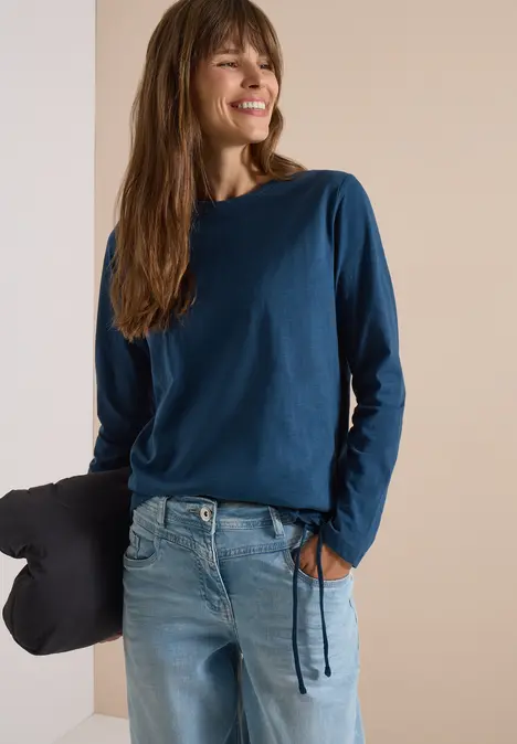 Eine Frau in blauem Langarmshirt und Jeans lehnt lächelnd an einer Wand, hält ein Kissen in der Hand.