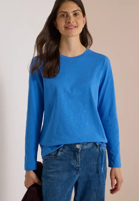Eine Frau steht vor hellem Hintergrund, trägt einen hellblauen Pullover und Jeans und lächelt.