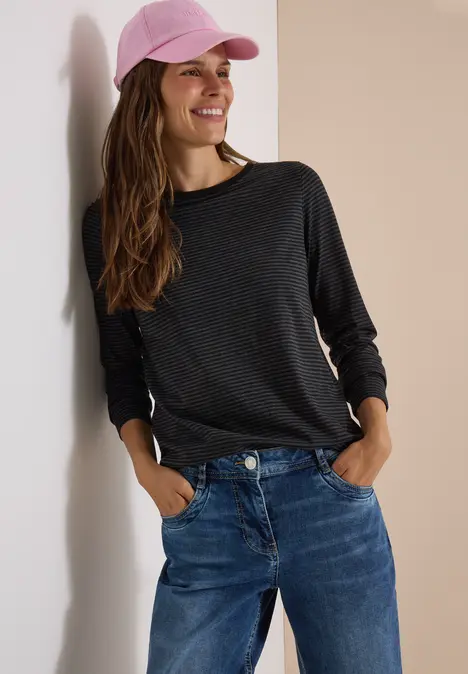 Frau lehnt an Wand, trägt schwarzen Langarmshirt, Jeans und rosa Baseballcap, lächelt.