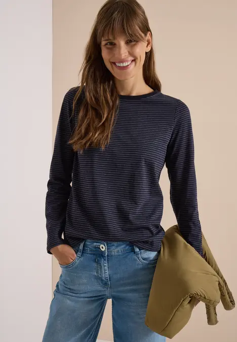 Frau in Jeans und gestreiftem Pullover hält Jacke, lächelt.