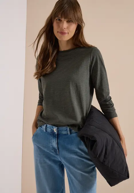 Junge Frau trägt grünes Langarmshirt und blaue Jeans, hält schwarze Jacke in der Hand.