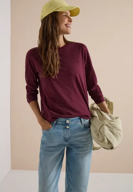 Frau trägt burgunderfarbenen Pullover, Jeans und grüne Baseballkappe, hält Tasche lässig über der Schulter.