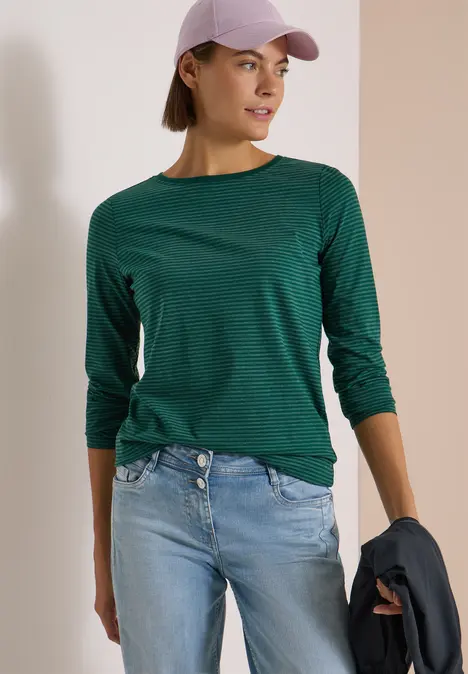 Frau mit grünem Langarmshirt, Jeans, Cap und Jacke in der Hand vor zweifarbigem Hintergrund.