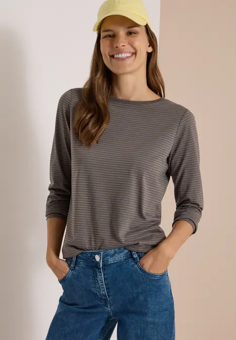 Eine Frau lächelt, trägt gestreiftes Langarmshirt, blaue Jeans und gelbe Baseballkappe.