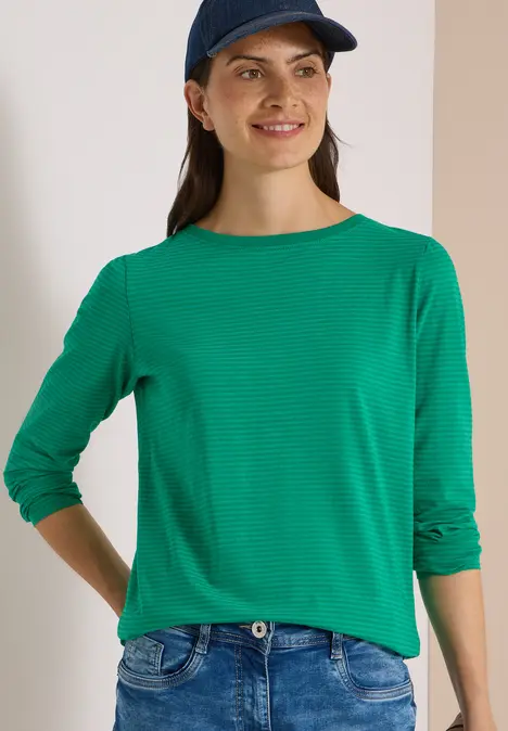 Junge Frau mit grüner Bluse und blauer Baseballkappe lächelt vor neutralem Hintergrund.