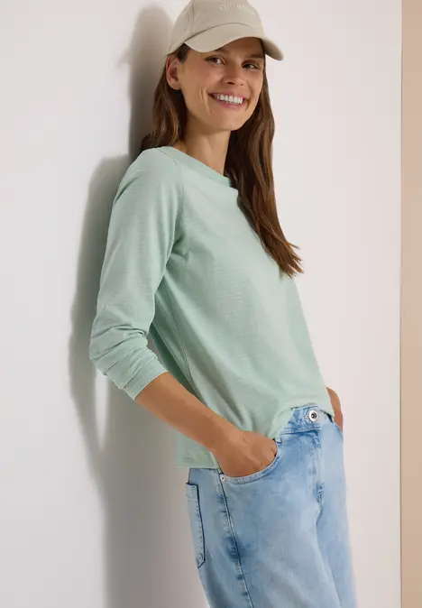 Eine Frau mit hellgrünem Langarmshirt, Jeans und Baseballcap lehnt lächelnd an einer Wand.