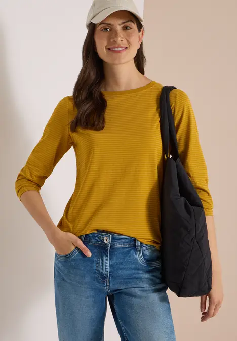 Junge Frau in gelbem Langarmshirt, Jeans, Baseballcap und schwarzer Umhängetasche lächelt.