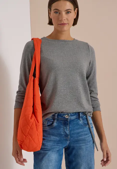 Eine Frau trägt einen grauen Pullover, blaue Jeans und eine orange Stepp-Tasche über der Schulter.
