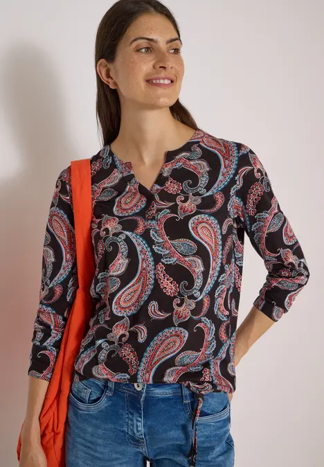 Frau trägt schwarze Bluse mit rotem Paisley-Muster, Jeans und rote Umhängetasche, lächelt entspannt.