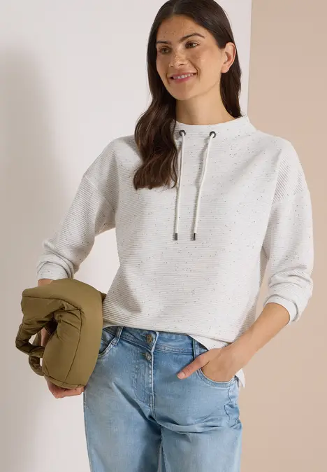 Junge Frau im weißen Hoodie und Jeans, die eine grüne Jacke hält und lächelt.