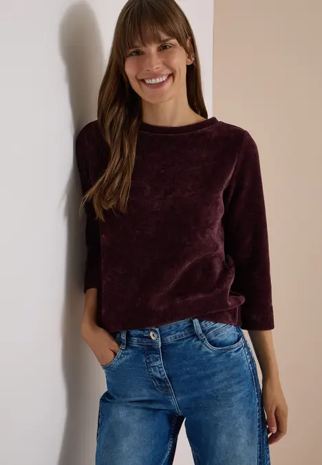 Junge Frau lehnt lächelnd an einer Wand, trägt lila Pullover und blaue Jeans.