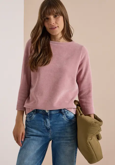 Frau in pinkem Pullover und Jeans, hält eine olivgrüne Jacke, stehend vor neutralem Hintergrund.