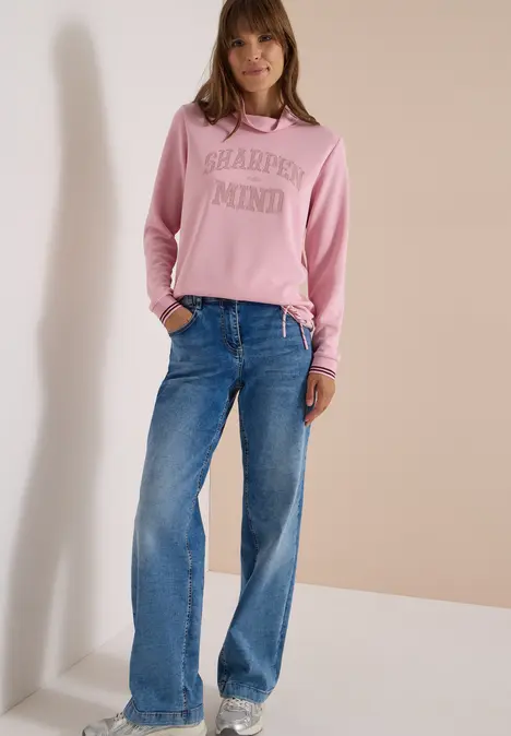 Eine Frau in rosa Kapuzenpullover und blauen Jeans vor neutralem Hintergrund.