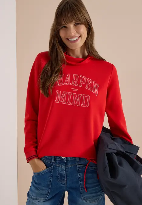 Eine lächelnde Frau im roten Sweatshirt 'OPEN MIND', in Jeans, hält eine Jacke vor neutralem Hintergrund.