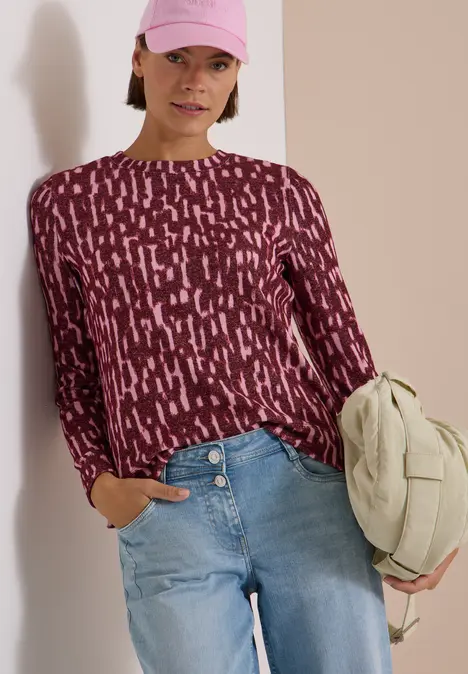 Eine Frau trägt einen bordeauxfarbenen gemusterten Pullover, blaue Jeans, rosa Cap und hält eine helle Tasche.