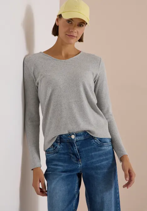 Femme portant un haut gris à manches longues, un jean bleu et une casquette jaune, debout sur fond clair.