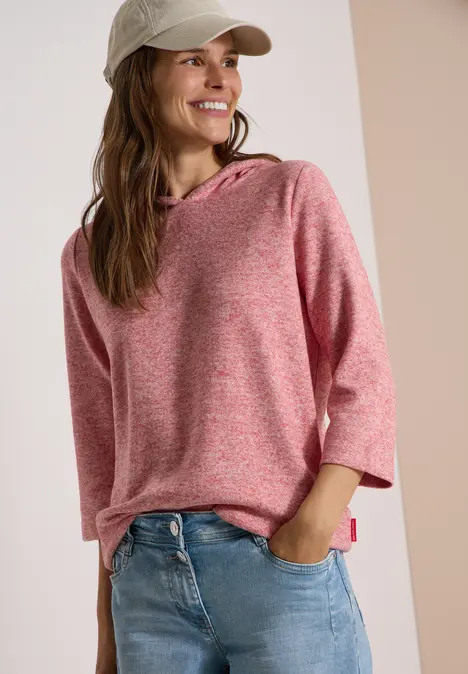 Uśmiechnięta kobieta w różowym hoodie z 3/4 rękawem, dżinsach i czapce.